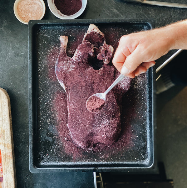 Raw fowl on sheet tray getting sprinkled with blueberry powder and salt